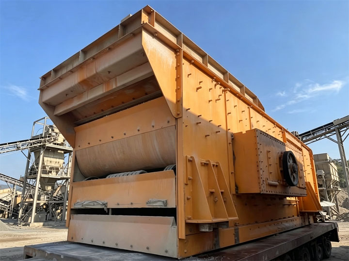 Massive industrial elliptical vibrating screen operating in an outdoor aggregate quarry site processing wet mineral materials.