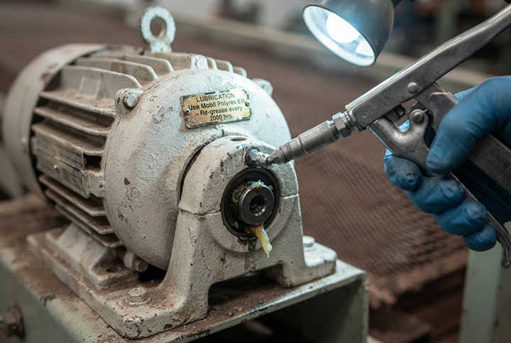 Extreme close-up of a maintenance technician in a blue glove using a grease gun to lubricate a heavy-duty vibration motor, with a lubrication instruction label clearly visible and fresh grease purging from the bearing housing.