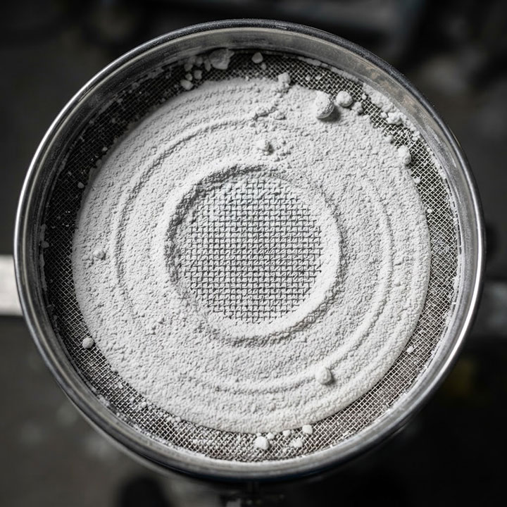An extreme close-up of an industrial vibrating screen mesh completely blinded and clogged with compacted fine white powder, showing a dense, solid cake.
