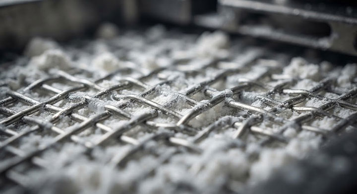 A macro close-up of a metal wire mesh from an industrial vibrating screen, showing clumps of moist white powder adhering to the wires and completely clogging the mesh openings.