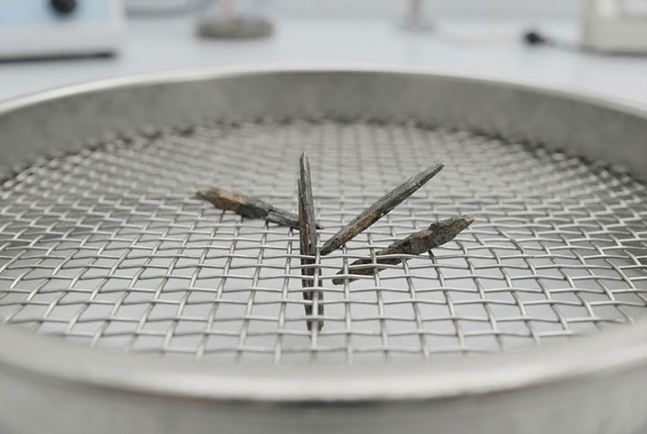 An extreme close-up macro photograph of a precision woven wire mesh test sieve, specifically illustrating mesh pegging where several elongated, needle-like mineral particles are wedged vertically and diagonally halfway through the square apertures.
