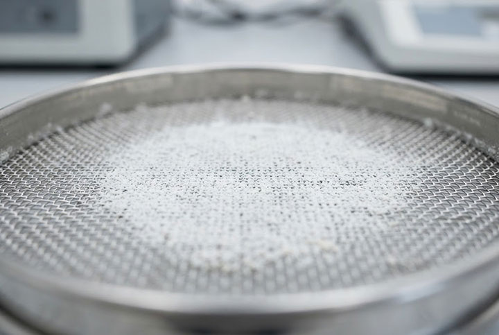 Macro close-up of sticky white pharmaceutical powder blinding a fine stainless steel wire mesh test sieve.