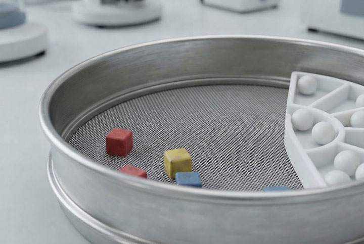 Macro close-up of colorful rubber and urethane cleaning cubes and a ball tray resting on the support grid beneath a stainless steel laboratory test sieve mesh.