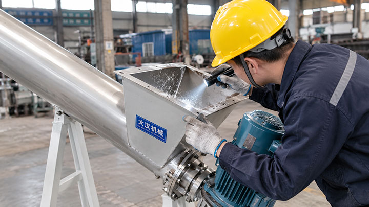 Industrial maintenance worker wearing a hard hat and gloves conducting a daily pre-operation inspection on a stainless steel screw powder conveyor, using a flashlight to check the material inlet for residual powder buildup.